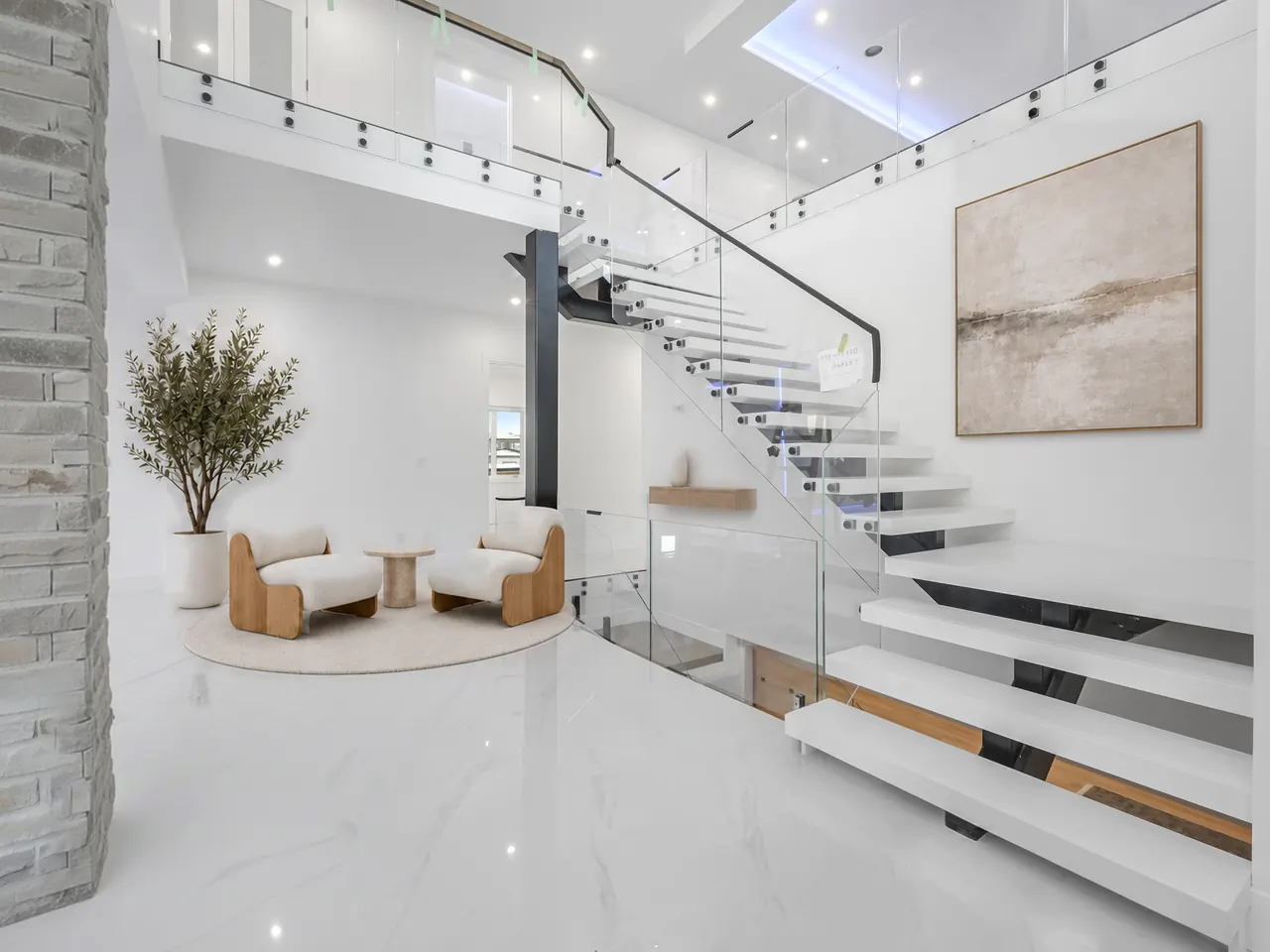 Floating staircase with glass railings in a double-height entry, virtually staged with two lounge chairs, an olive tree and an abstract canvas