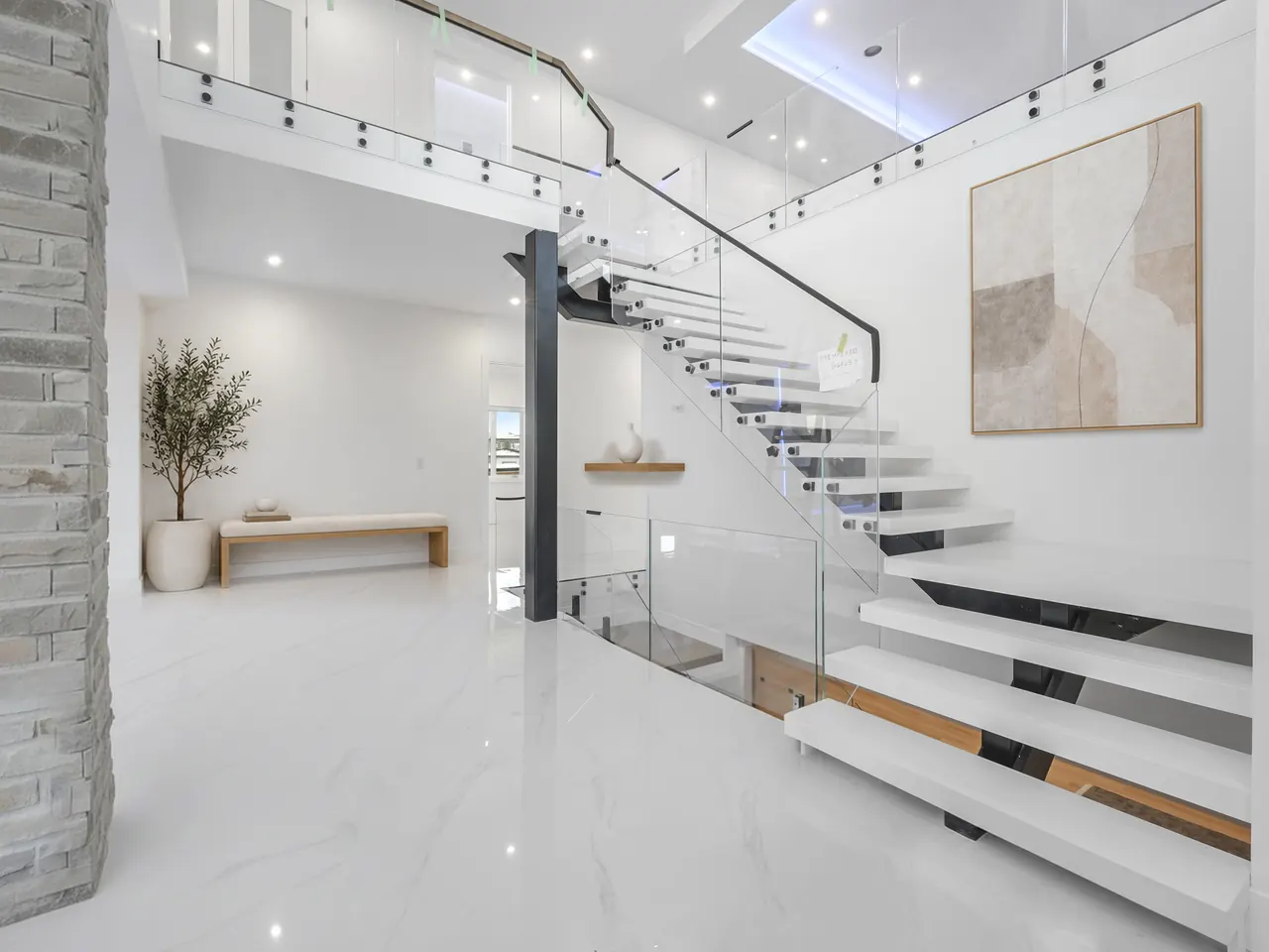 Floating staircase with glass railings in a double-height entry, virtually staged with an olive tree, an oak bench against the wall and an abstract canvas, leaving the way to the stairs clear